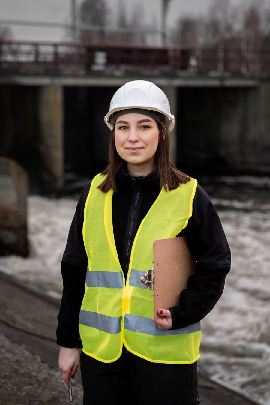 Ingeniera ambiental con casco, chaleco y carpeta junto a una represa
